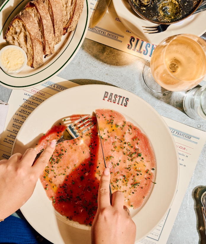 An overhead view of hands cutting into a large salmon dish on a Pastis restaurant plate, with bread, wine, and menus on the table.