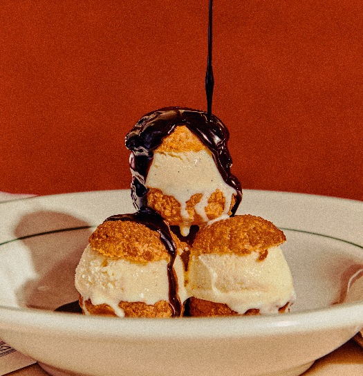 Three cream puffs filled with vanilla ice cream stacked in a bowl with chocolate sauce drizzled on top.