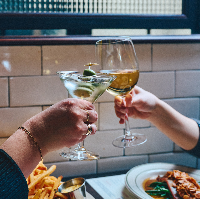 Two hands toasting with cocktail glasses over a restaurant table with food including fries and plated dishes