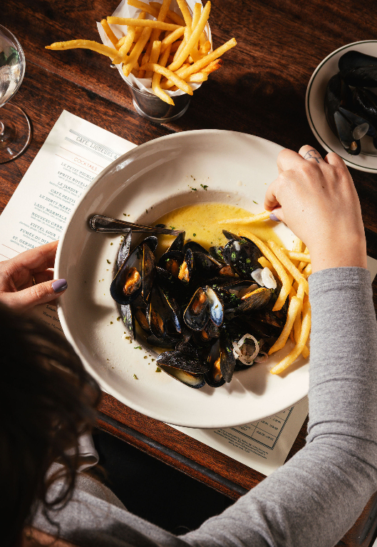 Person eating moules frites - black mussels with french fries and yellow sauce on white plate at wooden table