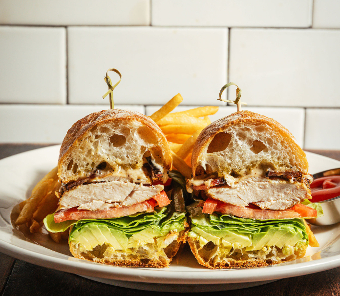 Chicken sandwich cut in half on ciabatta bread with lettuce, tomato, and avocado, served with french fries on white plate