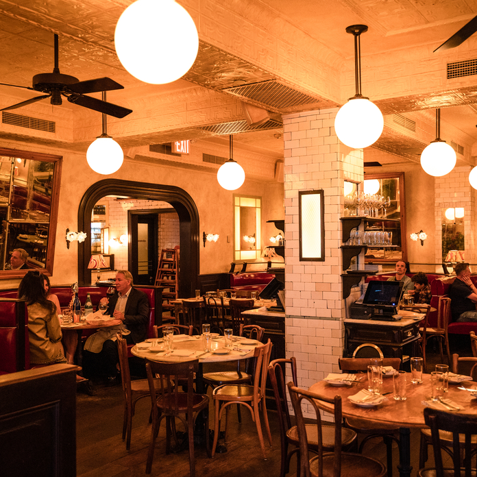 Cozy, warmly lit restaurant with round tables, brick wall, globe lights, patrons dining, and a vintage, inviting ambiance.