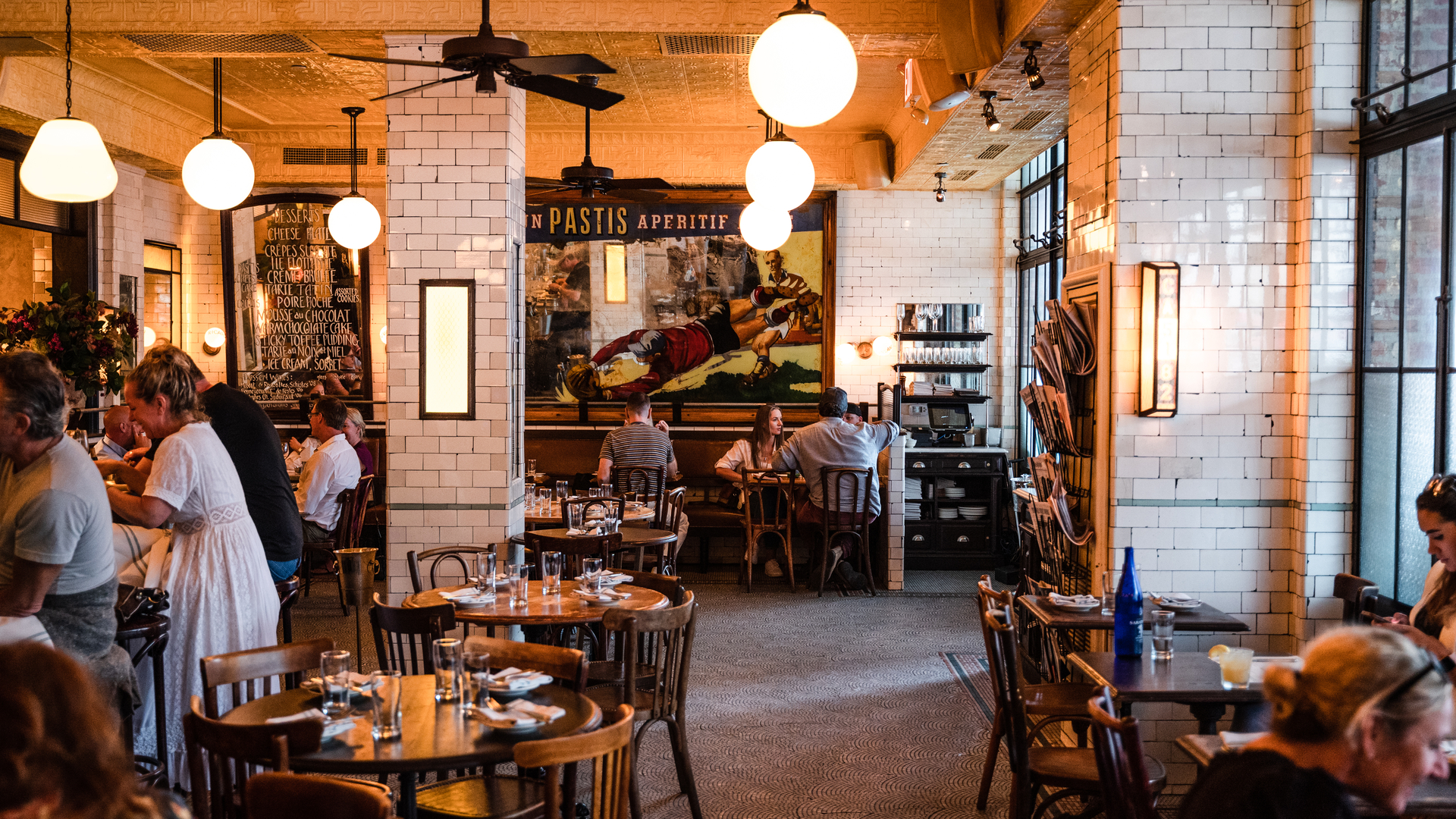 Warm, cozy restaurant interior with round tables, brick walls, large windows, and patrons dining or waiting.