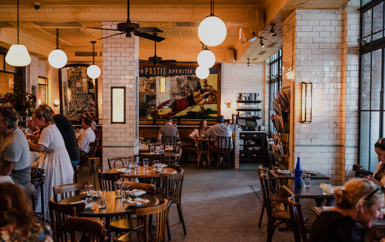 A cozy restaurant interior with white-tiled walls, wooden tables, chairs, and patrons dining. Hanging lights and a large mural on the back wall.