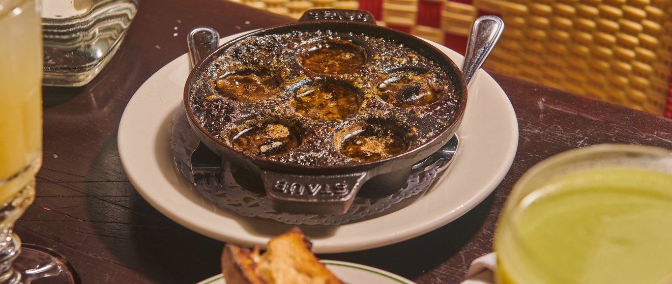 Gourmet escargot dish in a black pan, served with bread and a green beverage, on a wooden table with a woven mat background.